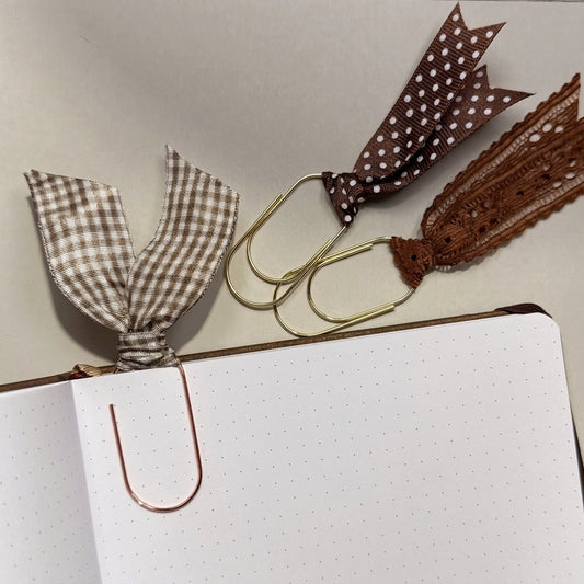 Three decorative paperclips styled on a notebook: a brown and beige gingham ribbon clip, a brown polka-dot ribbon clip with a lace tassel, and a rose gold metal U-shaped clip holding an open page.