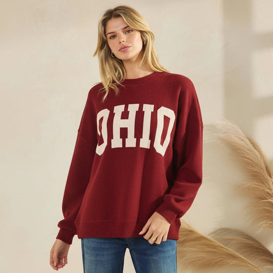A woman wearing a red-colored crewneck sweatshirt with 'OHIO' printed in cream block letters across the chest, paired with blue jeans.