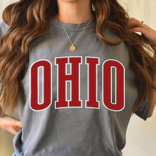 A woman wearing a gray graphic t-shirt with 'OHIO' printed in red and white varsity letters, paired with blue jeans.