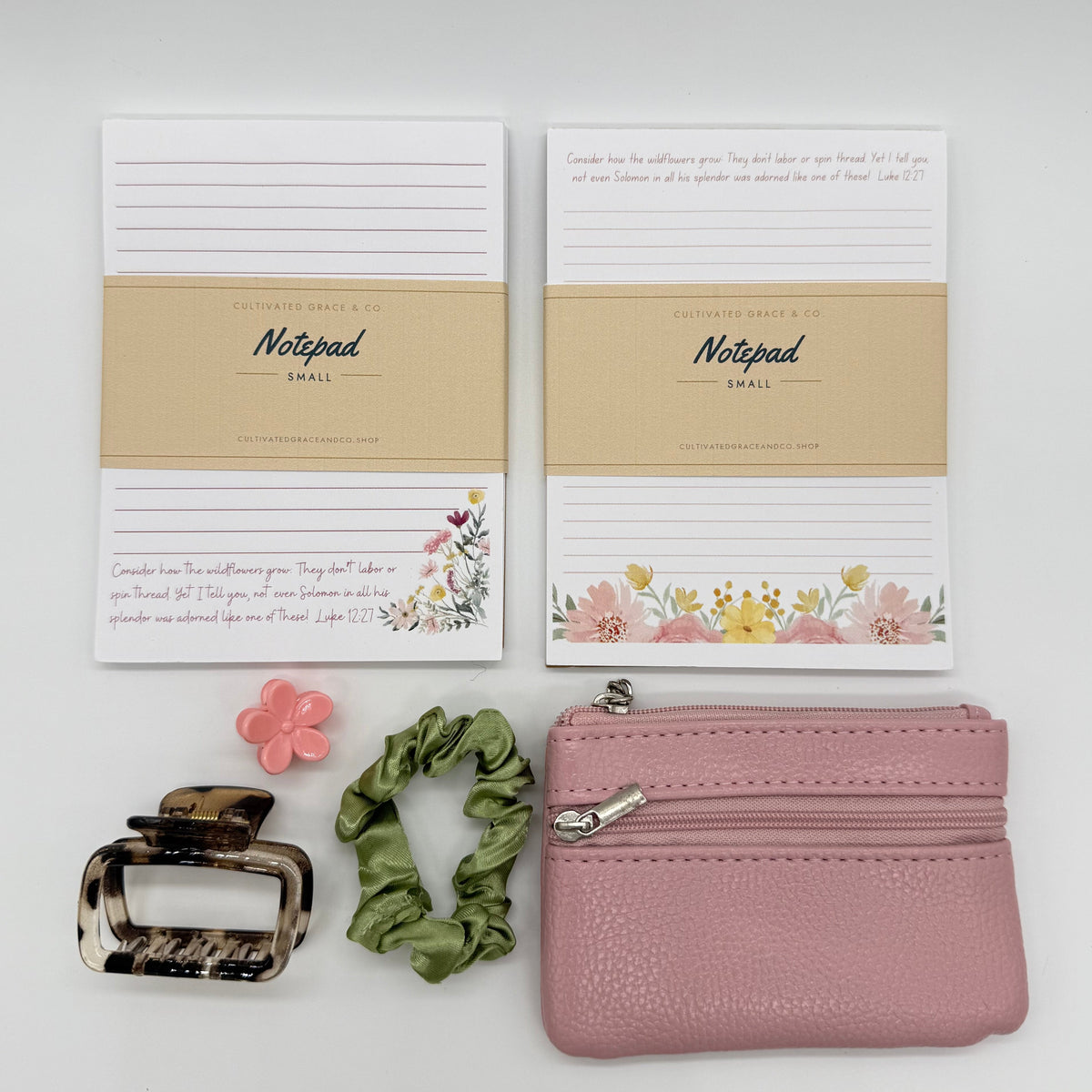 A flat-lay product arrangement on a light gray background featuring two Notepad (Eiffel) memo pads, a pink textured zippered pouch, a green scrunchie, a tortoiseshell hair clip, and a small pink flower accessory.