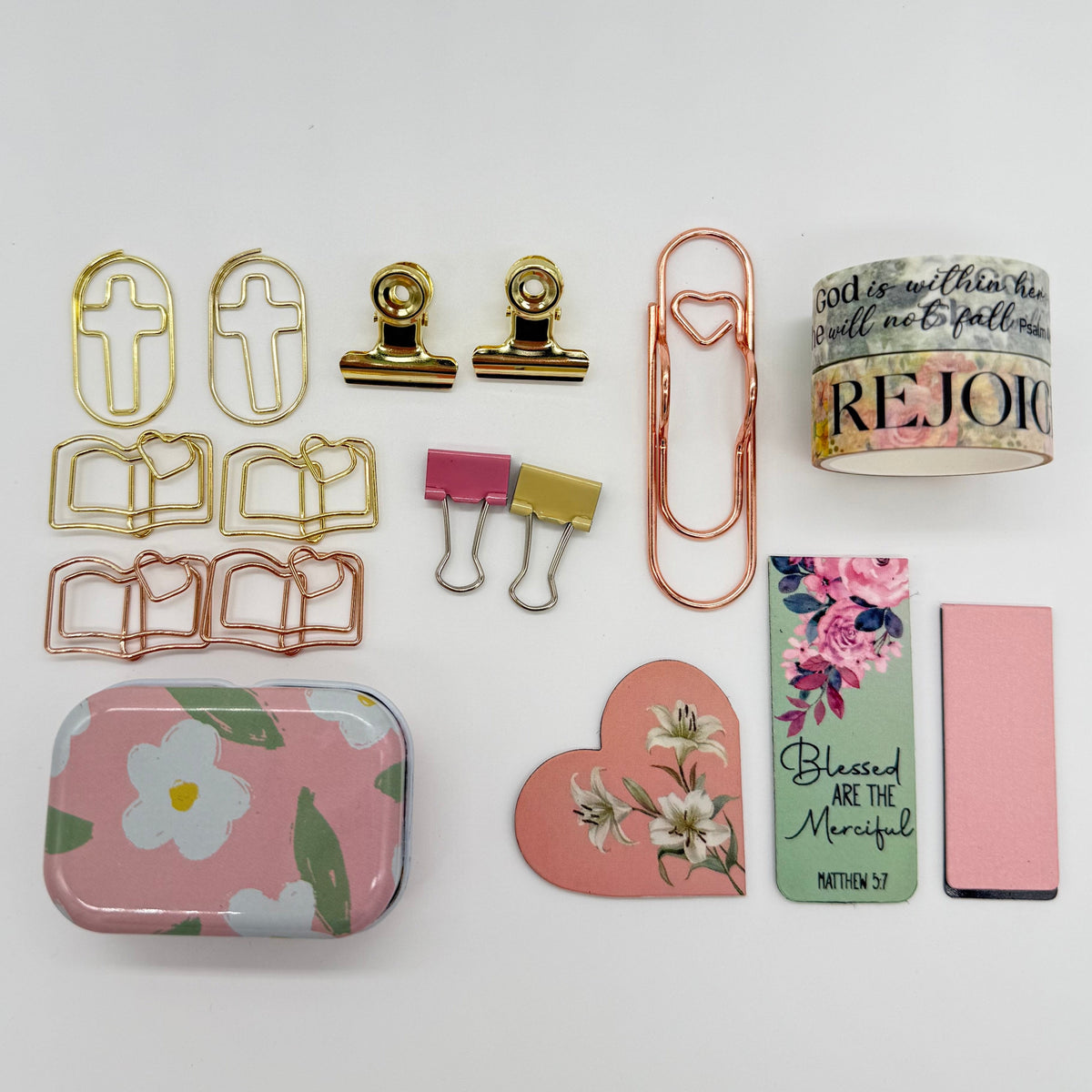 A flat-lay collection of stationery and desk accessories on a white background, including a pink floral tin, gold and rose gold paperclips, a pink and gold binder clip, a floral heart sticker, a green and pink card, a pink rectangular item, and a roll of washi tape.