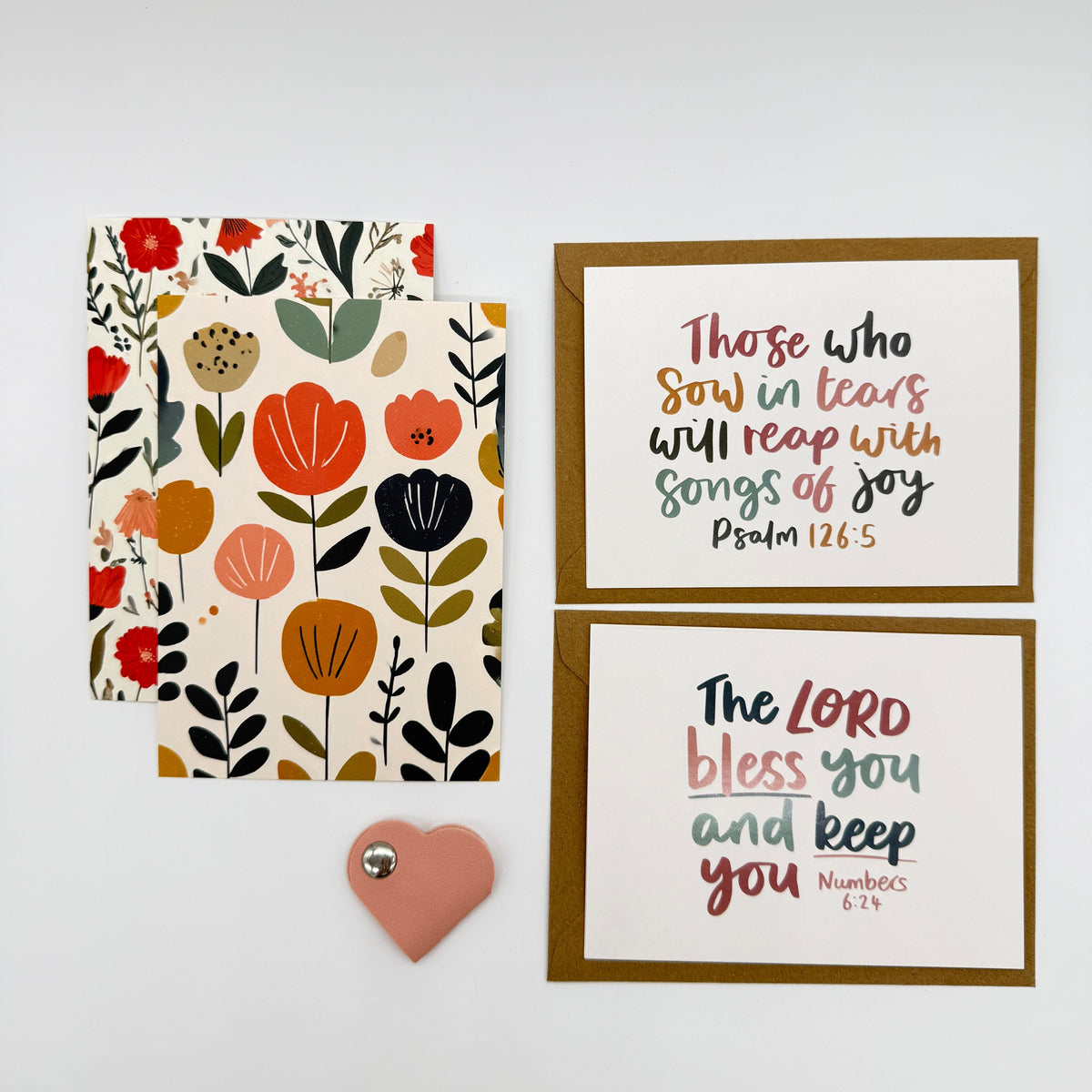 A flat-lay of a coordinated stationery set on a light gray background, including a floral patterned envelope, a small pink heart-shaped tag, and two framed art prints with inspirational Bible verses.