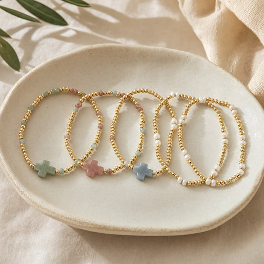 Assortment of gold beaded stretch bracelets with sage, rose, and blue cross charms and pearl accents on a cream dish