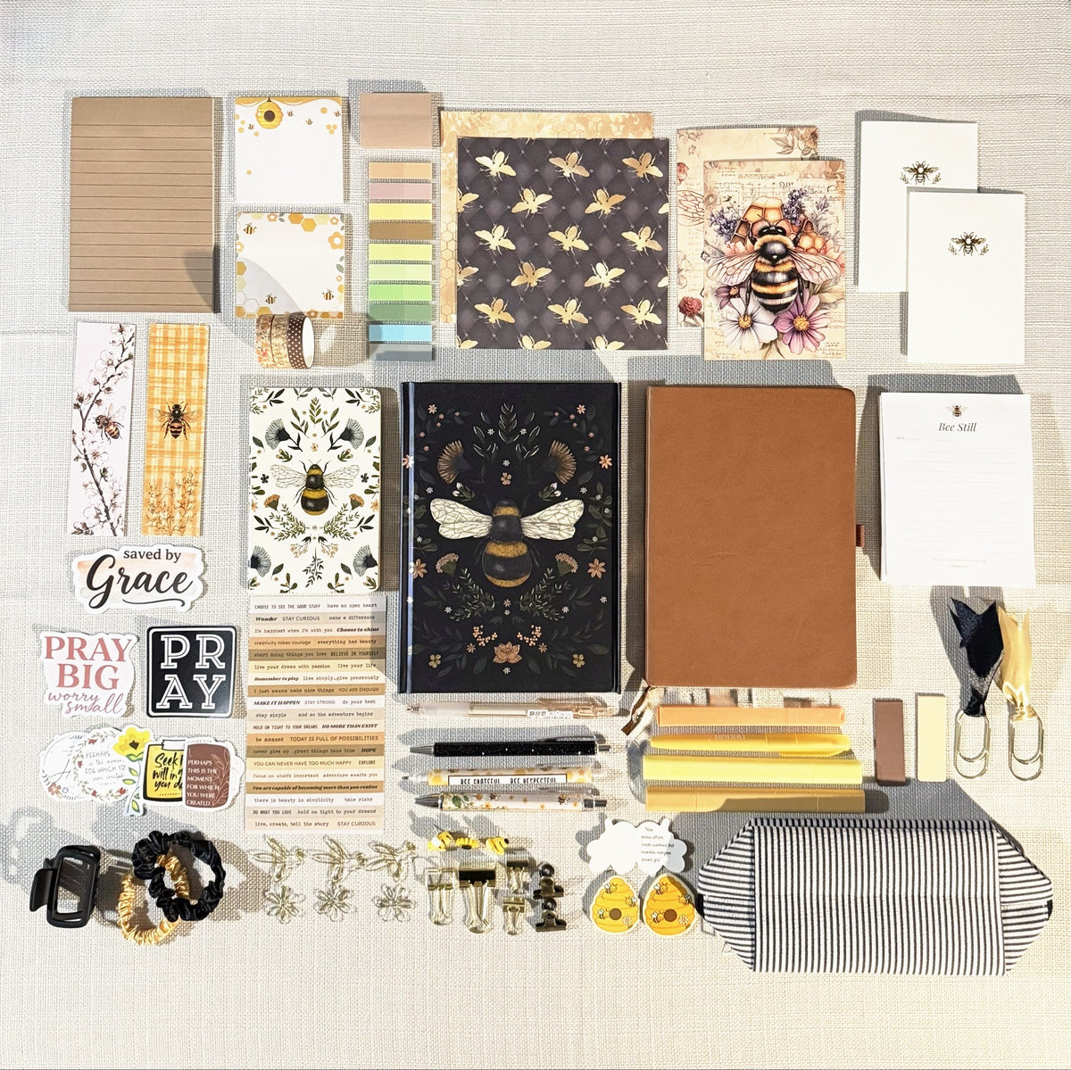Honey Bee Box contents arranged flat: black botanical bee journal, brown journal, mini bee notebook, Bee Still notepad, bee postcards and notecards, pens, highlighters, bookmarks, clips, stickers, zip pouch and accessories. Colors and individual items vary.