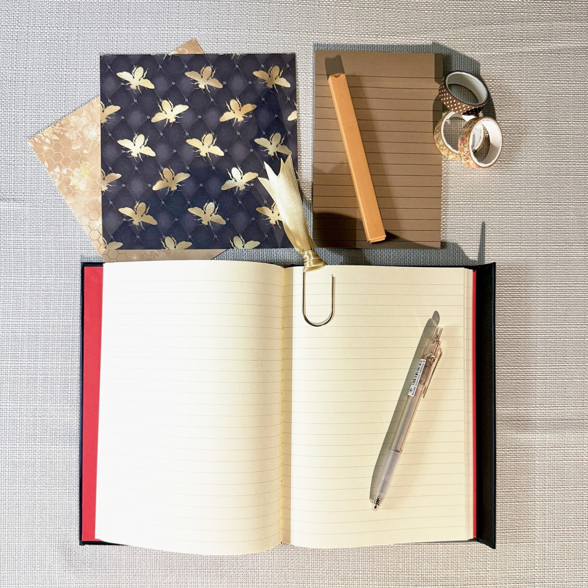 Open lined journal with a ribbon paperclip bookmark, pen, sticky notes and bee-themed styling; items vary.