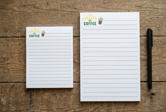 Two Sunshine and Coffee notepads on a rustic wood desk with a pen